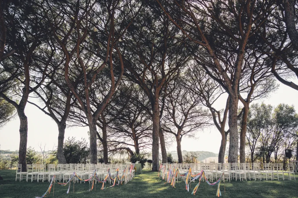 Outdoor wedding ceremony with rows of white chairs decorated with colorful ribbons, under a canopy of tall trees.