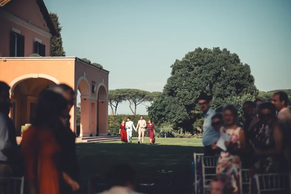 A couple walks down the aisle outdoors, flanked by two people, with wedding guests seated on either side and a large house in the background.