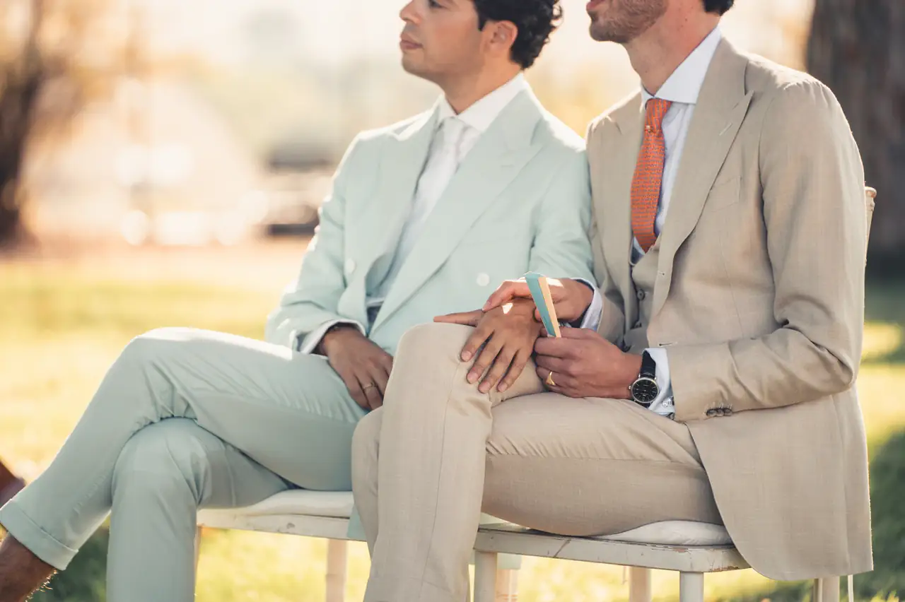 Two individuals are sitting close together outdoors, both dressed in light-colored clothing, one mint green and the other beige, and holding hands while sitting on a bench.