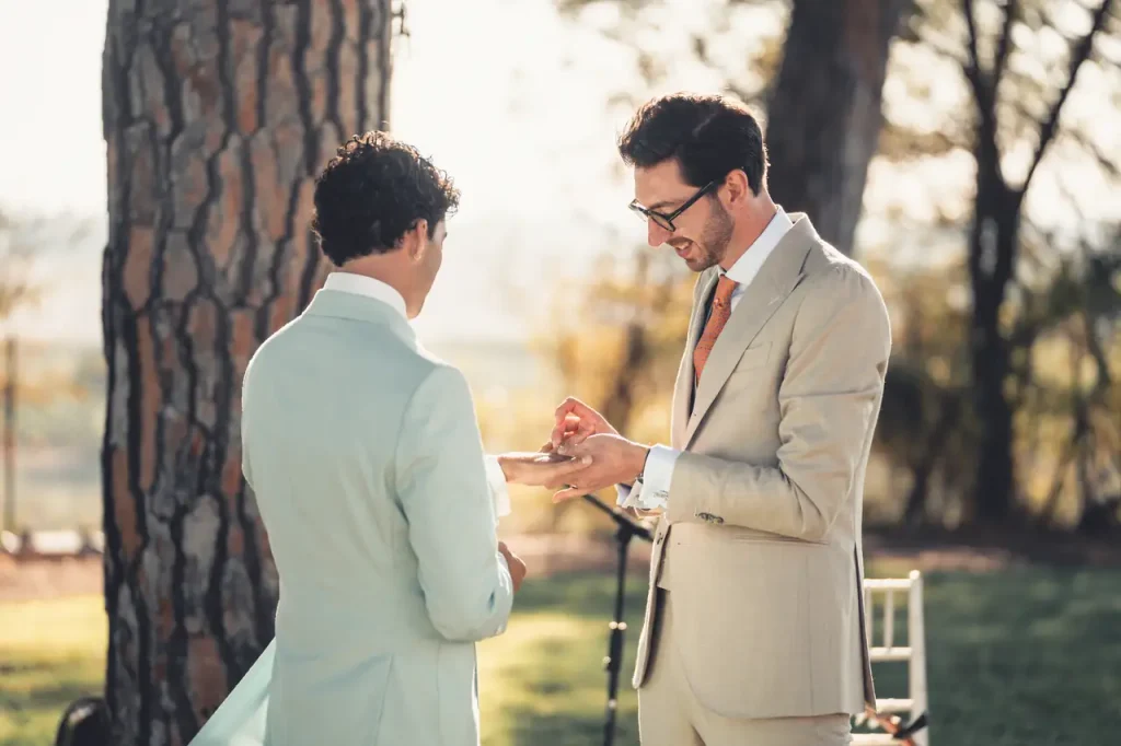 Two men in suits stand outside and exchange rings during a ceremony. One is holding a ring while the other is looking at it. Trees can be seen in the background.