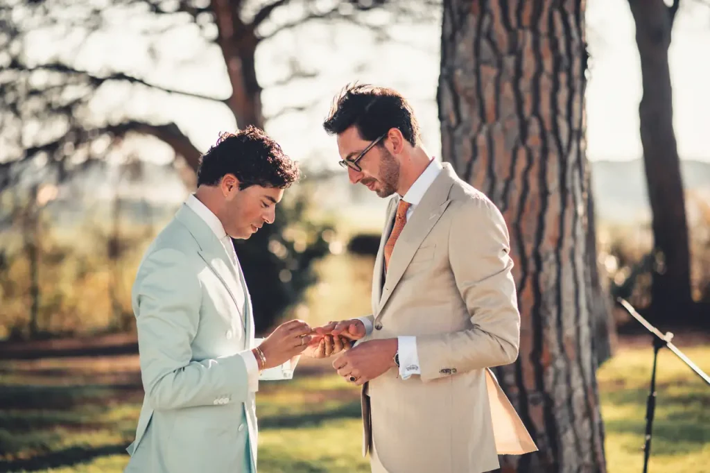 Two men in suits exchange rings outdoors during a wedding ceremony, surrounded by trees and natural light.