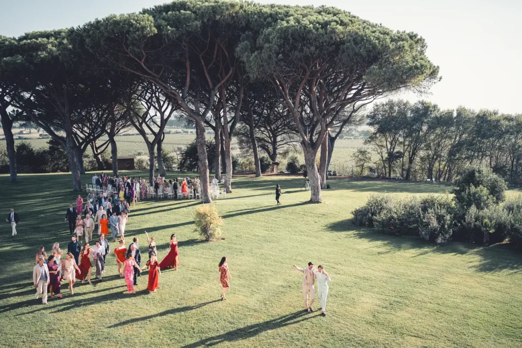A large group of people in colorful clothing gather under tall trees in a vast grassy field on a sunny day.