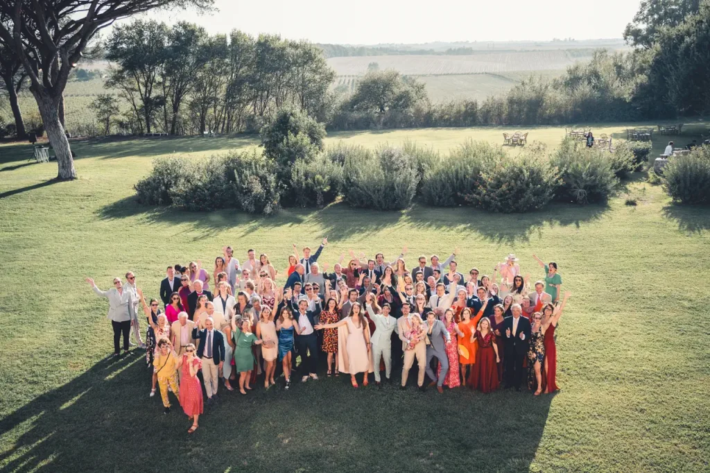A large group of people dressed in formal and casual attire pose together on a grassy lawn, with trees and a field in the distance in the background.