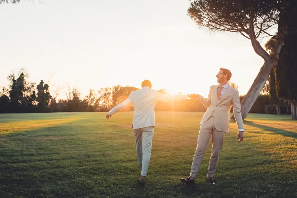 Villa alberico: two people in light-colored clothing dance joyfully on a grassy field at sunset, with trees in the background.