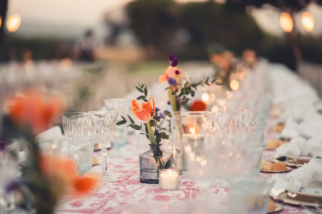 A long table elegantly set, decorated with glasses, small vases of flowers and lit candles in a serene outdoor setting.