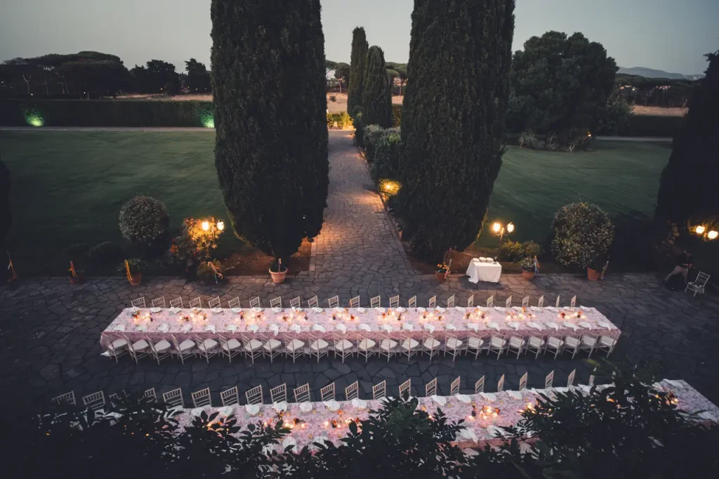 An outdoor dining set with two long tables covered with white cloth and surrounded by chairs. Tall trees and manicured lawns are visible in the background, with soft evening light illuminating the area.