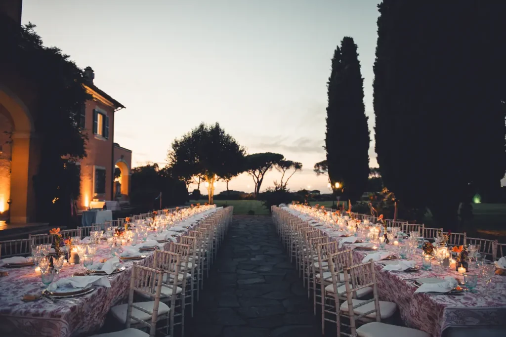 Villa alberico's outdoor evening setting includes long tables adorned with white tablecloths and chairs, lit by candles. The backdrop of majestic trees and the villa's charming architecture are bathed in warm sunset hues, creating an enchanting atmosphere.