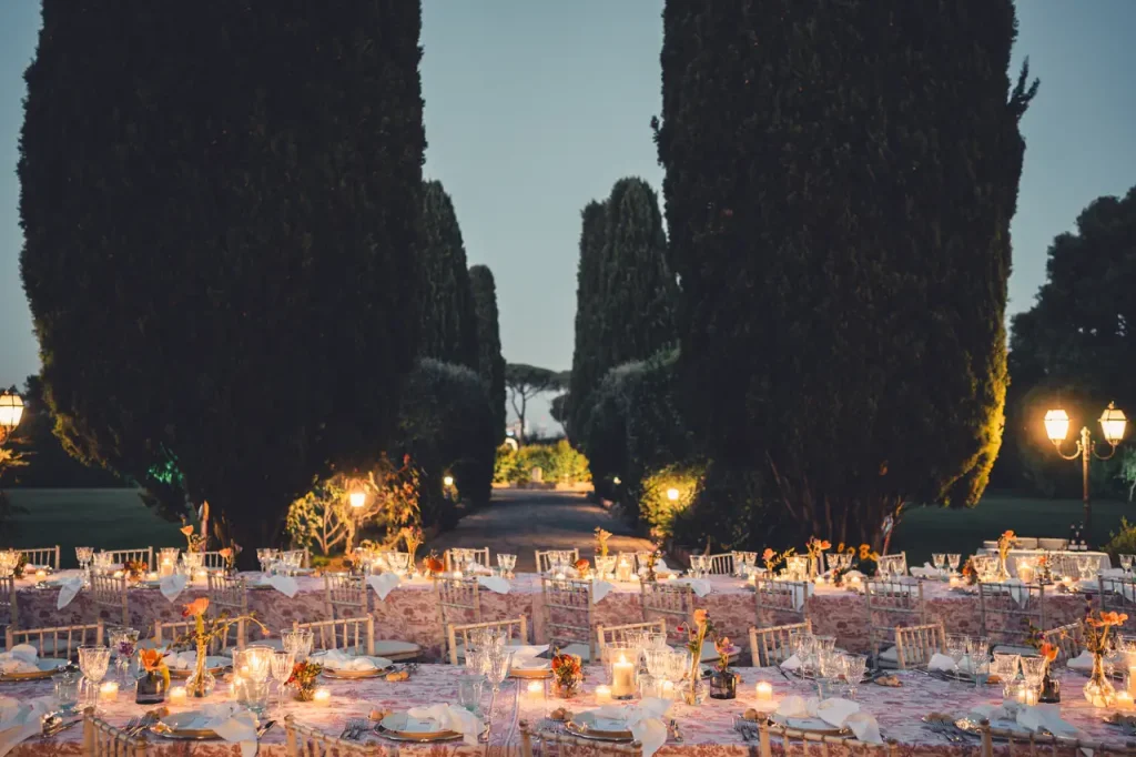 An evening banquet is set up outdoors at villa alberico among the tall hedges, with lighted tables and chairs leading to a distant path.