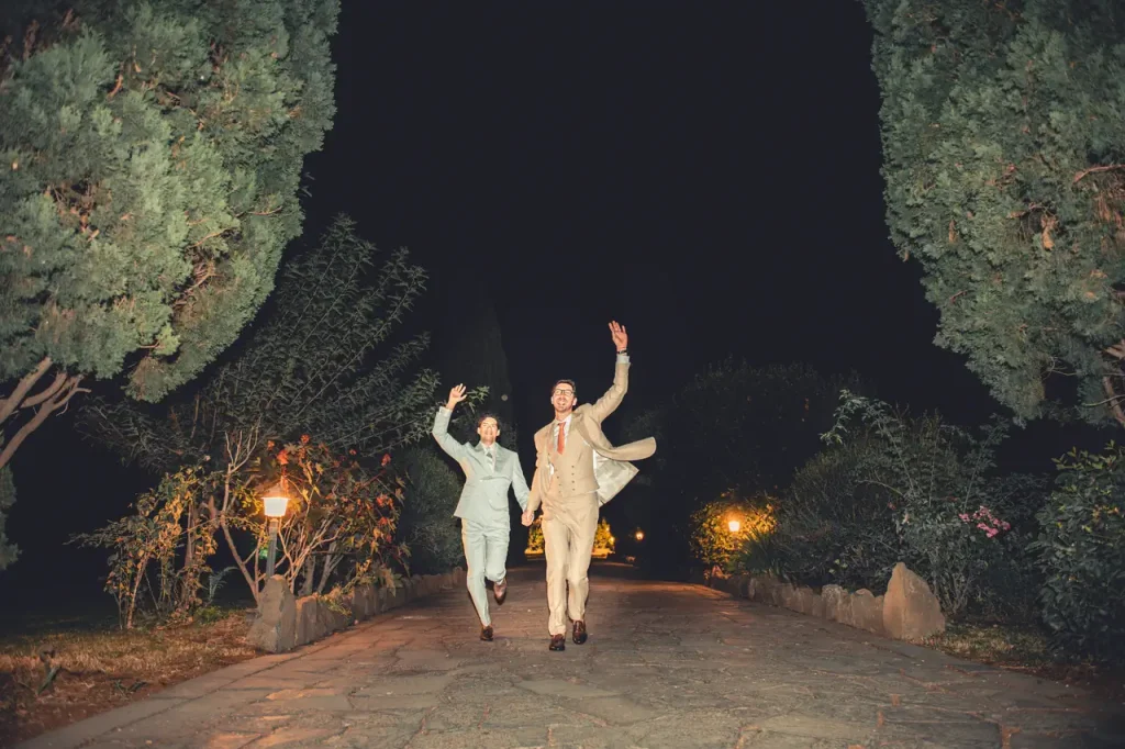 The bride and groom, in formal attire, run hand in hand on a stone path at night, waving with wide smiles. The path is lined with trees and lit by warm lights.