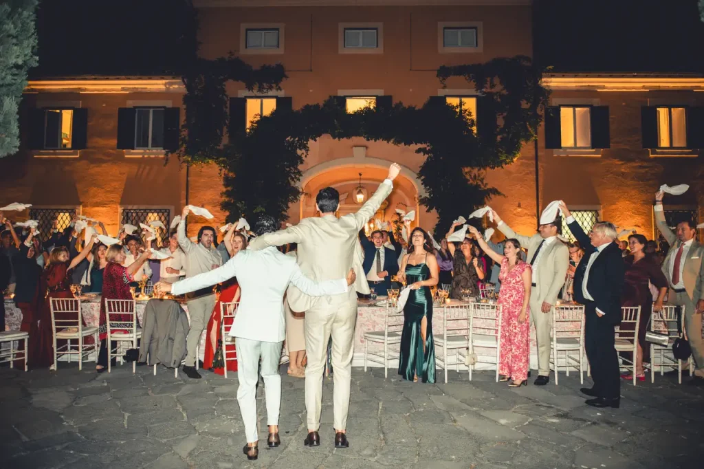 A joyful crowd waves napkins as formally dressed grooms stand with arms raised in front of a lighted building during an outdoor evening event.