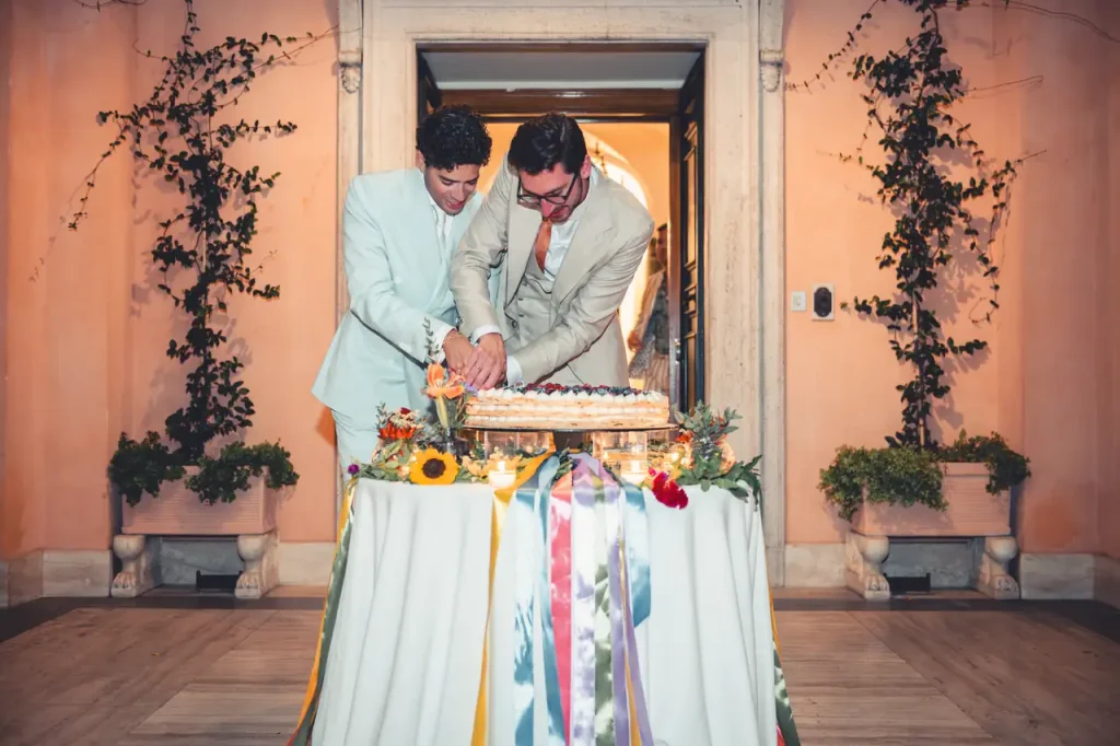 Two men in suits, the bride and groom, cut a wedding cake together on a table decorated with colorful ribbons and flowers, standing at a gateway decorated with greenery.