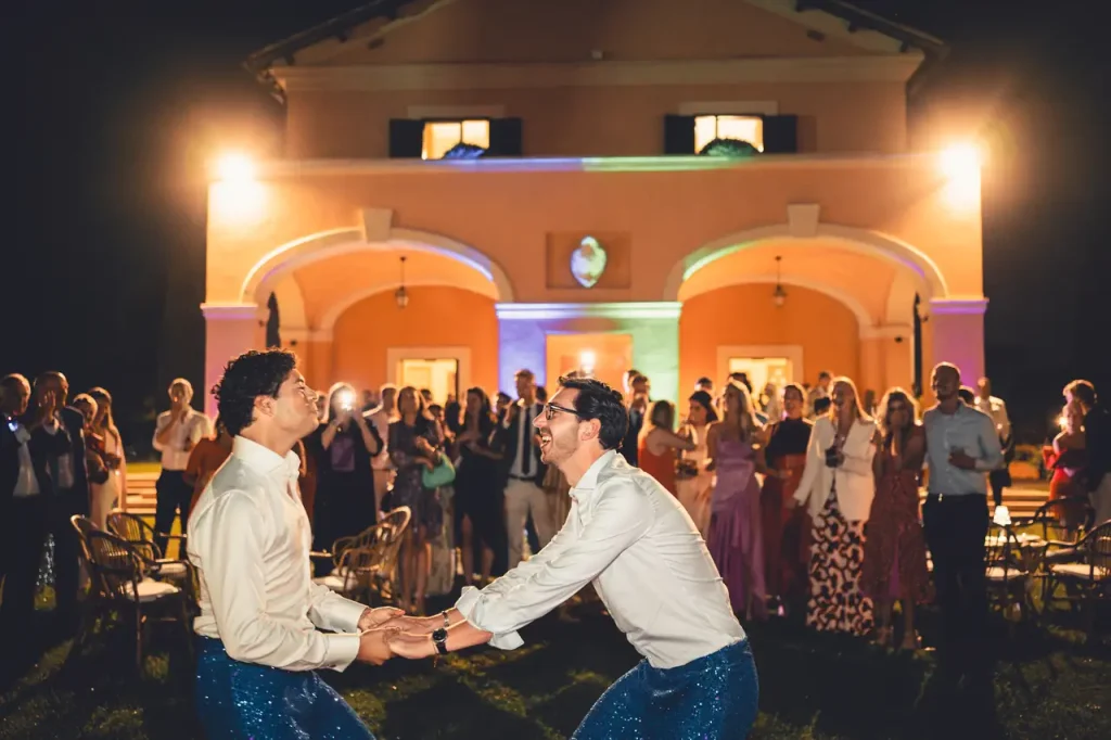 Two people hold hands and dance in front of a well-lit building at night, with a gathered crowd watching in the background; it is clear that the bride and groom are the center of attention.