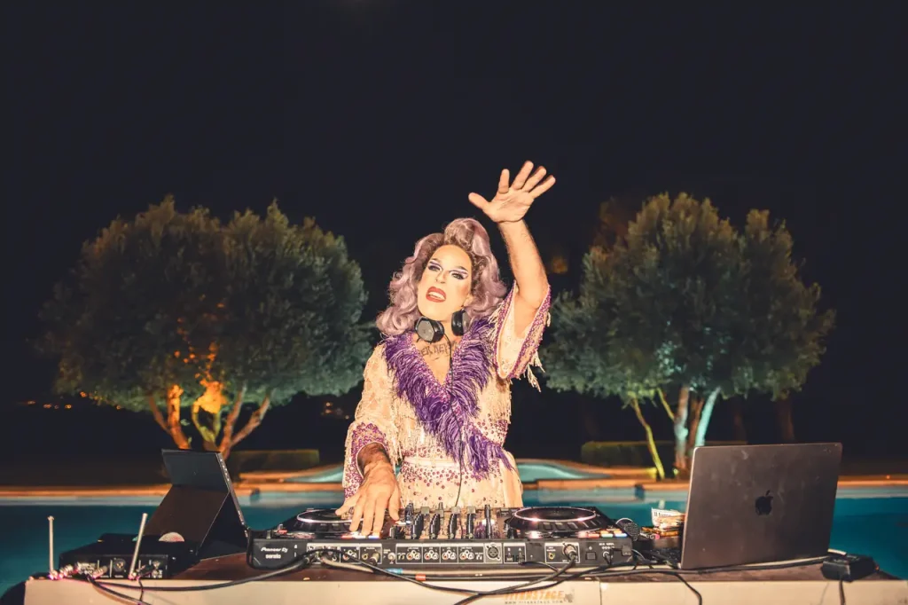 A dj in colorful clothes, with wig and makeup, performs, raising a hand, with equipment and two laptops on the table, against the backdrop of night trees.
