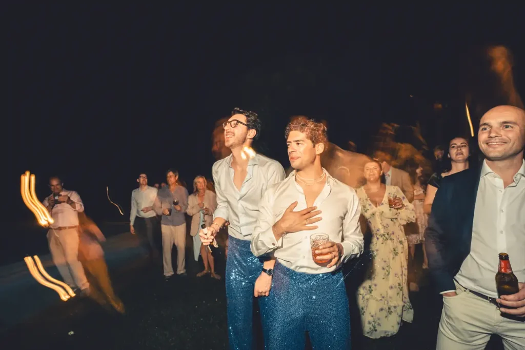 Two men in matching blue sequin pants, the bride and groom, hold drinks, surrounded by a group of people at an outdoor night event.