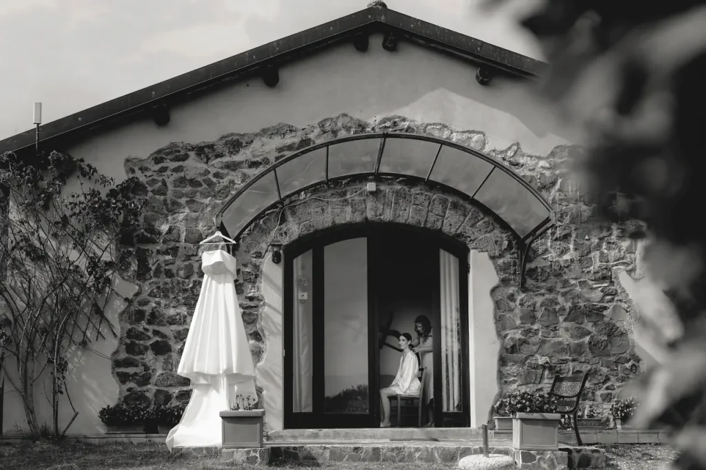 A stone building with an arched entrance, casale di polline exudes rustic charm. A wedding dress hangs gracefully outside, while a person is visible through the doorway, sitting in silent contemplation.