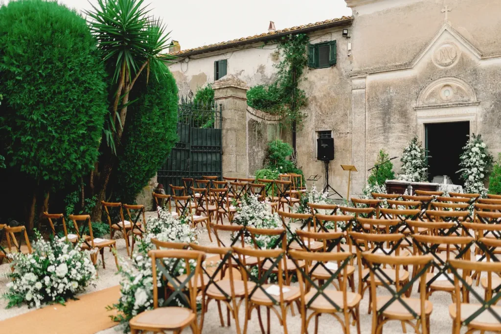 In the rustic charm of a courtyard, rows of wooden chairs and vibrant floral decorations await guests. The historic church of our lady of the most holy rosary stands gracefully in the background, adding an air of timeless elegance to the wedding's outdoor setting.