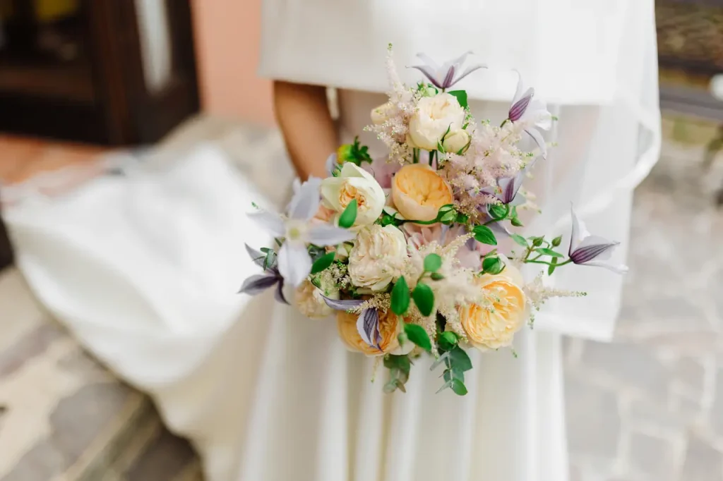 A bride in a white dress holds a bouquet of pastel flowers, including yellow roses and purple details, standing on a stone path.