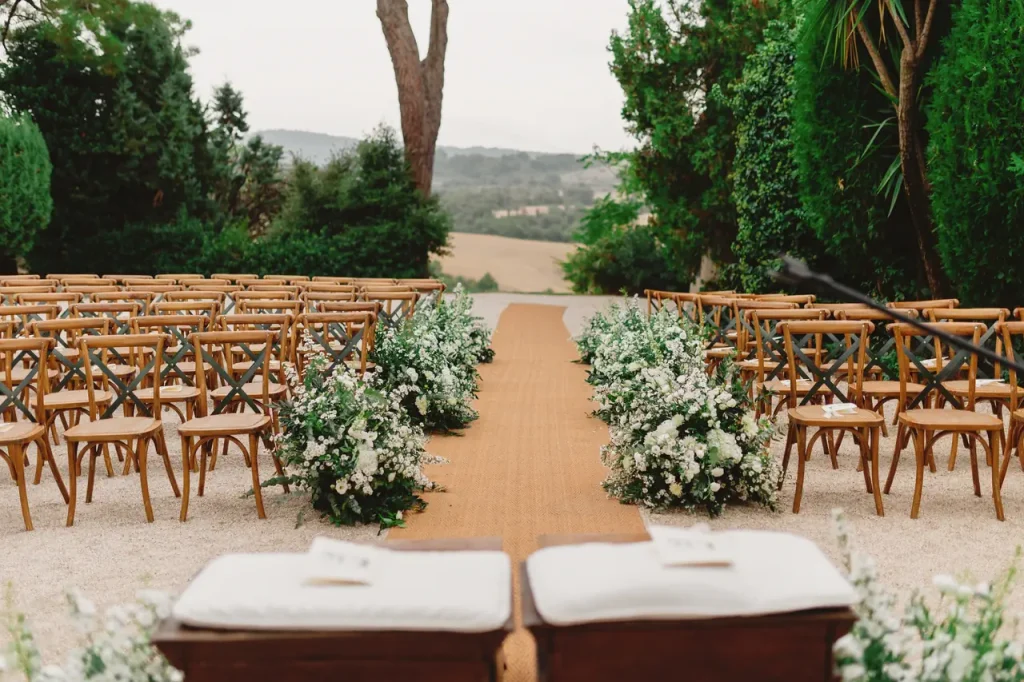 Outdoor wedding ceremony with rows of wooden chairs, a beige carpet down the aisle and white flower arrangements, all set in a lush garden.