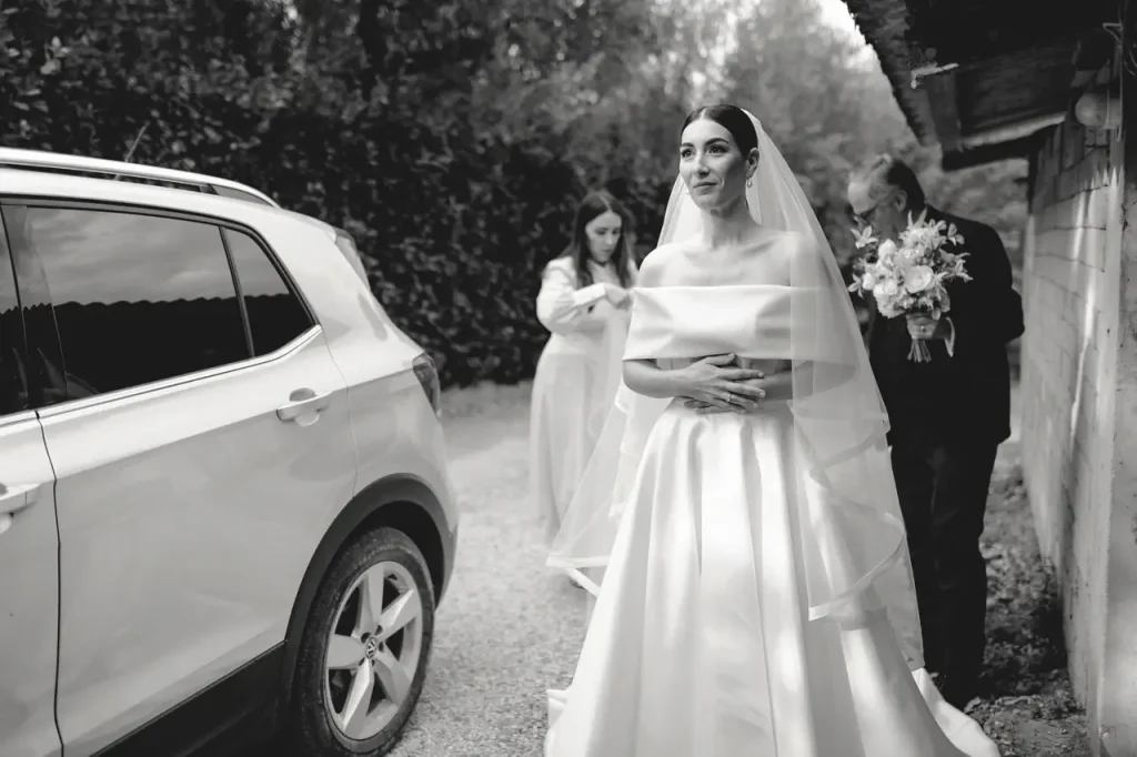 A bride in a white wedding dress and veil is standing next to a car, accompanied by two people holding flowers.