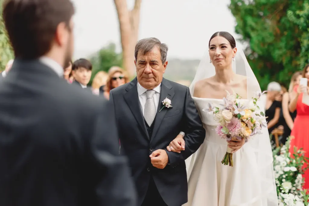 A bride in a white dress holds a bouquet as she walks down the aisle with an older man in a suit. Guests are seated in the background.