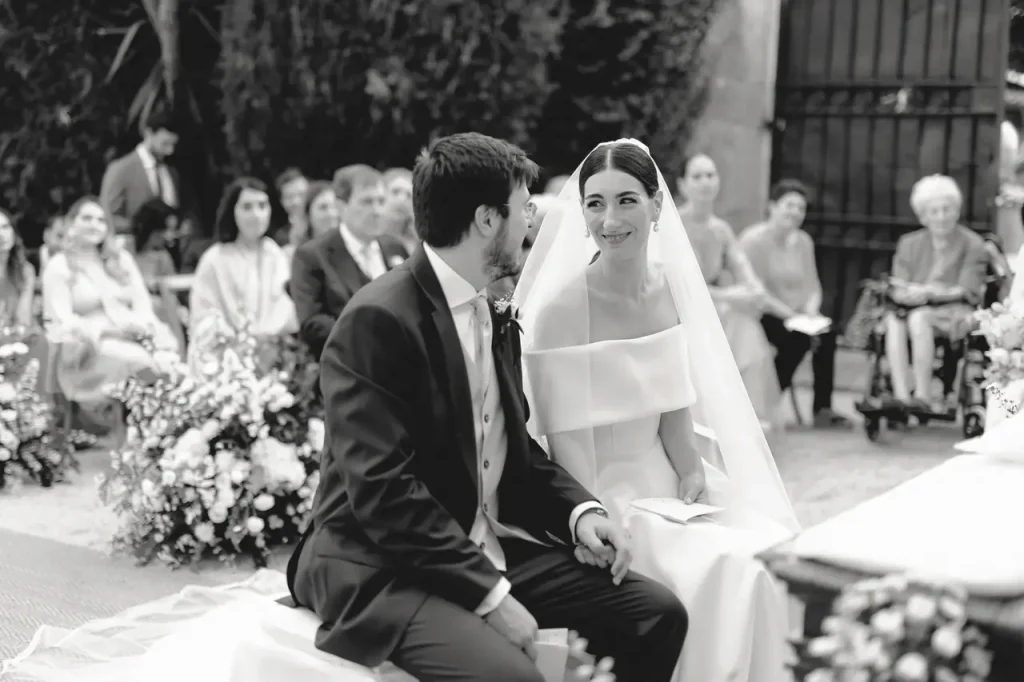 The bride and groom sit next to each other during an outdoor wedding ceremony, surrounded by seated guests and flower arrangements.