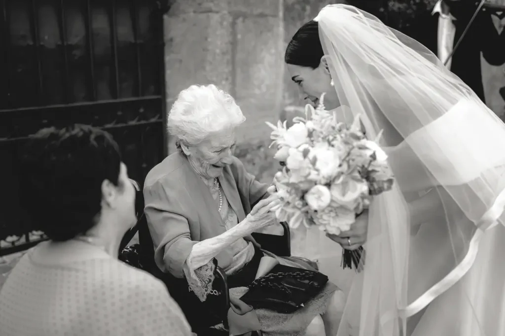 A bride with veil and bouquet in hand greets an elderly woman sitting next to another person outdoors.