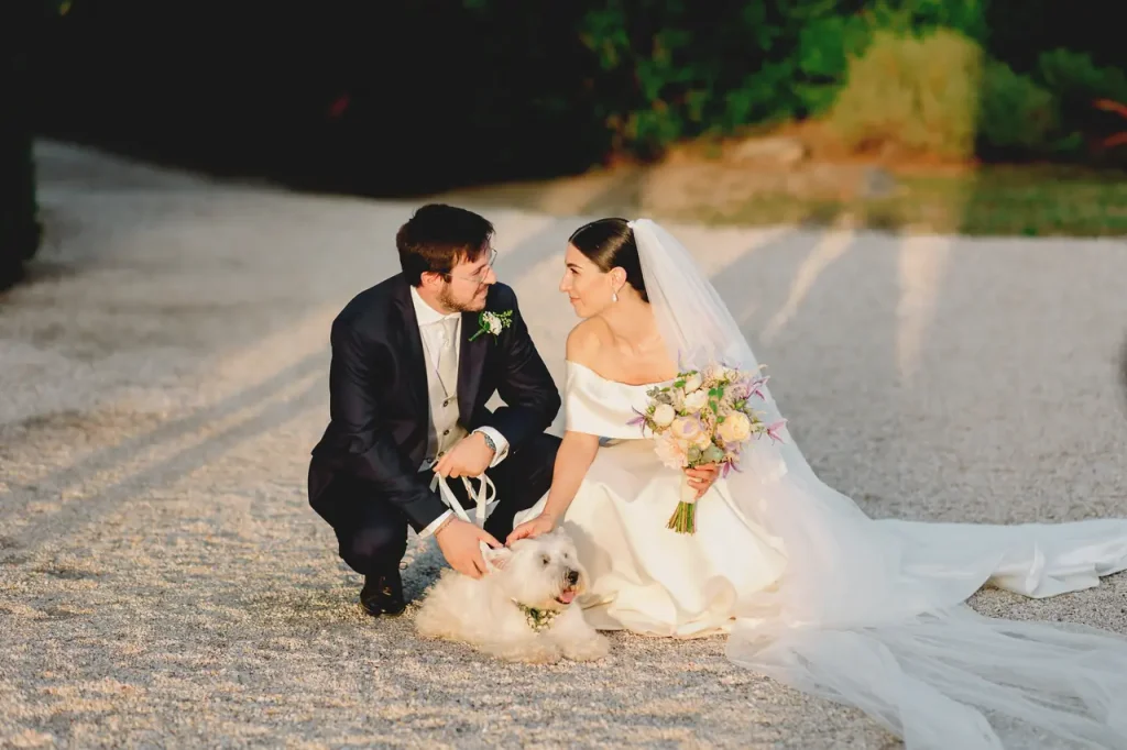 The newlyweds are sitting on a gravel path with a white dog. The bride is holding a bouquet and both are dressed in wedding dresses.