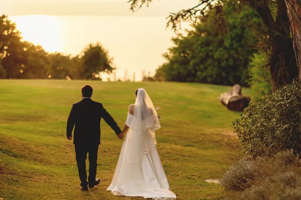 A bride and groom walk hand in hand on a grassy field near a lake at sunset.