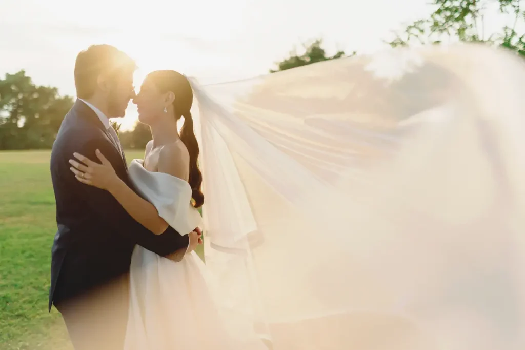 A groom and bride embrace outdoors, illuminated by the sun. The bride's veil floats in the breeze.