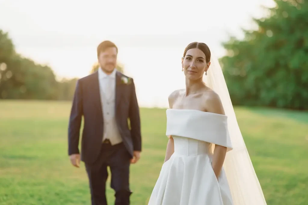 A bride in a off-the-shoulder dress poses outdoors with the groom in the background on a sunny day.