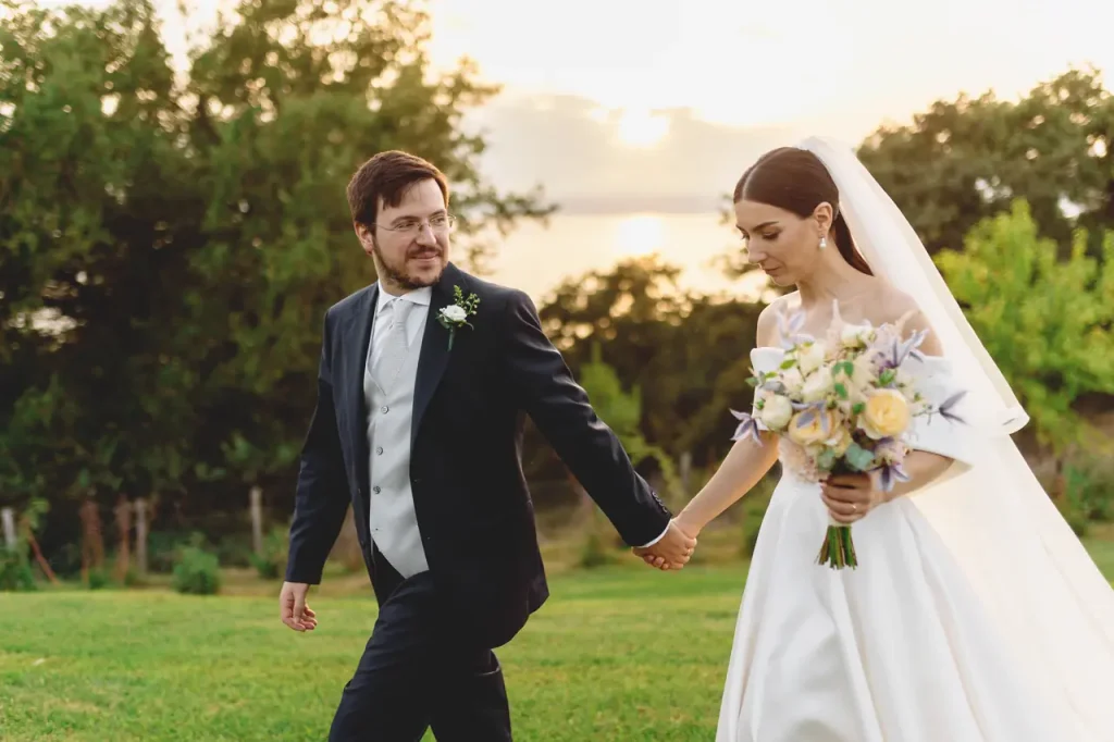 A bride and groom hold hands as they walk on a meadow, surrounded by trees. The bride wears a white dress and holds a bouquet, while the groom wears a dark suit.
