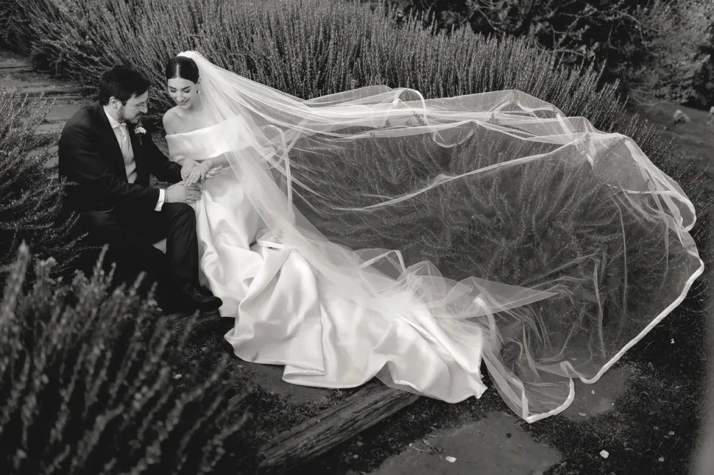 A bride and groom sit on a path, surrounded by grass and plants. The bride's long veil is spread beside them. The image is in black and white.