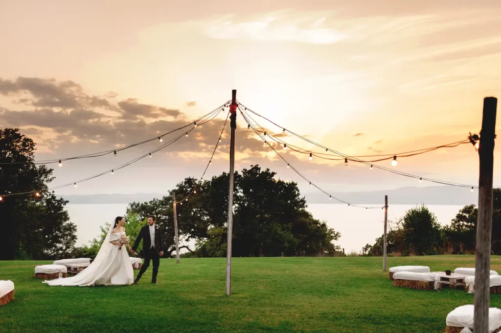 A couple in wedding dresses walk through a grassy field with string lights overhead at sunset. Trees and a body of water are in the background.