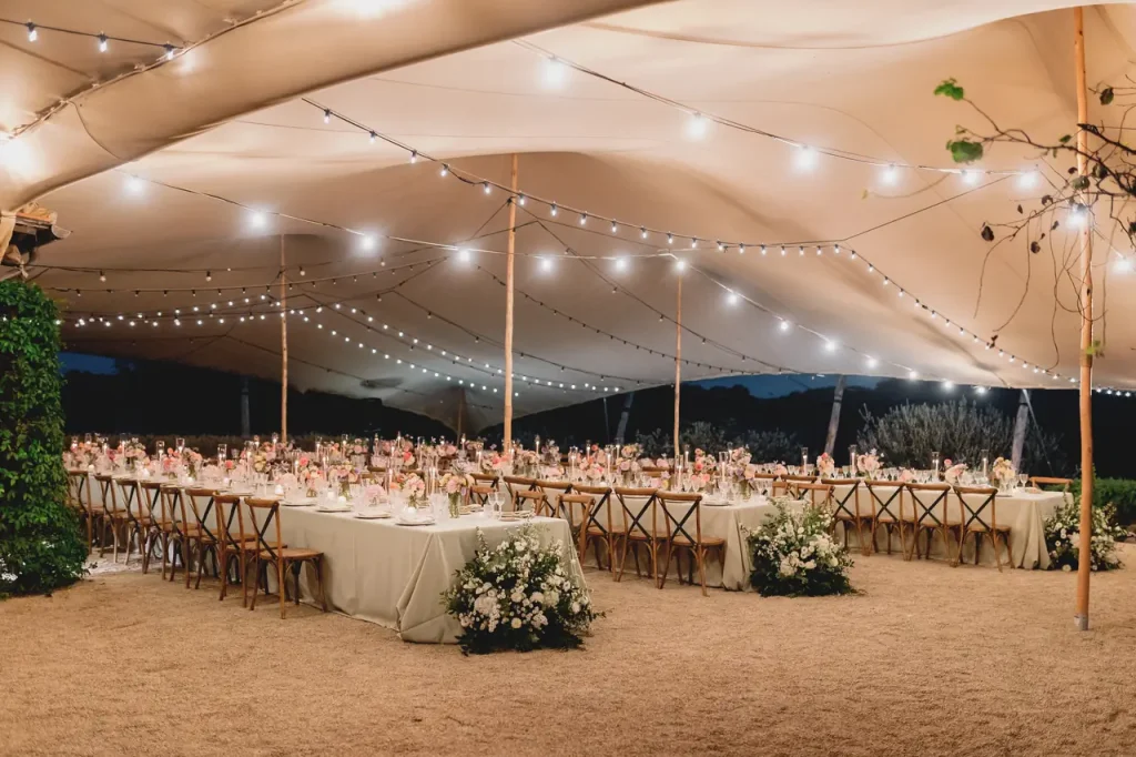 Outdoor wedding reception under a tent with string lights, long tables set with floral arrangements and wooden chairs on a sandy ground.