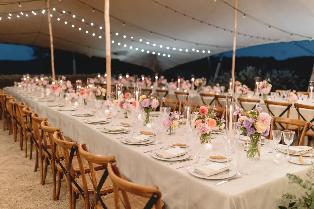 A long banquet table elegantly set under a curtain with string lights, floral centerpieces, candles, and equipment with plates and glasses.