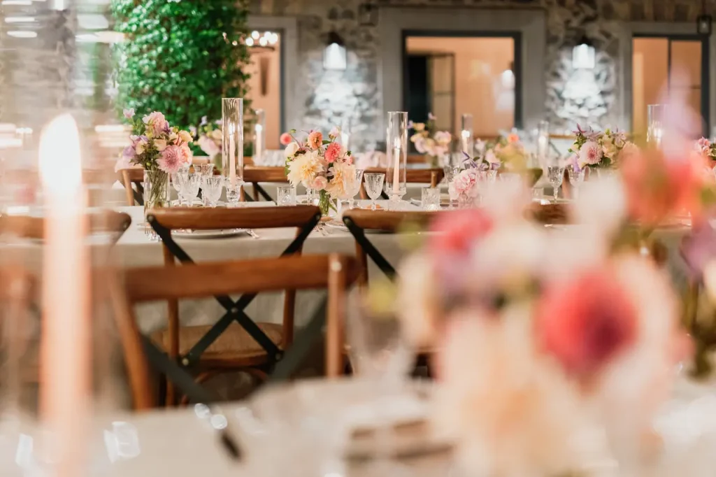 Set up for outdoor dinner with wooden chairs and tables decorated with floral centerpieces and candles, against a stone wall.