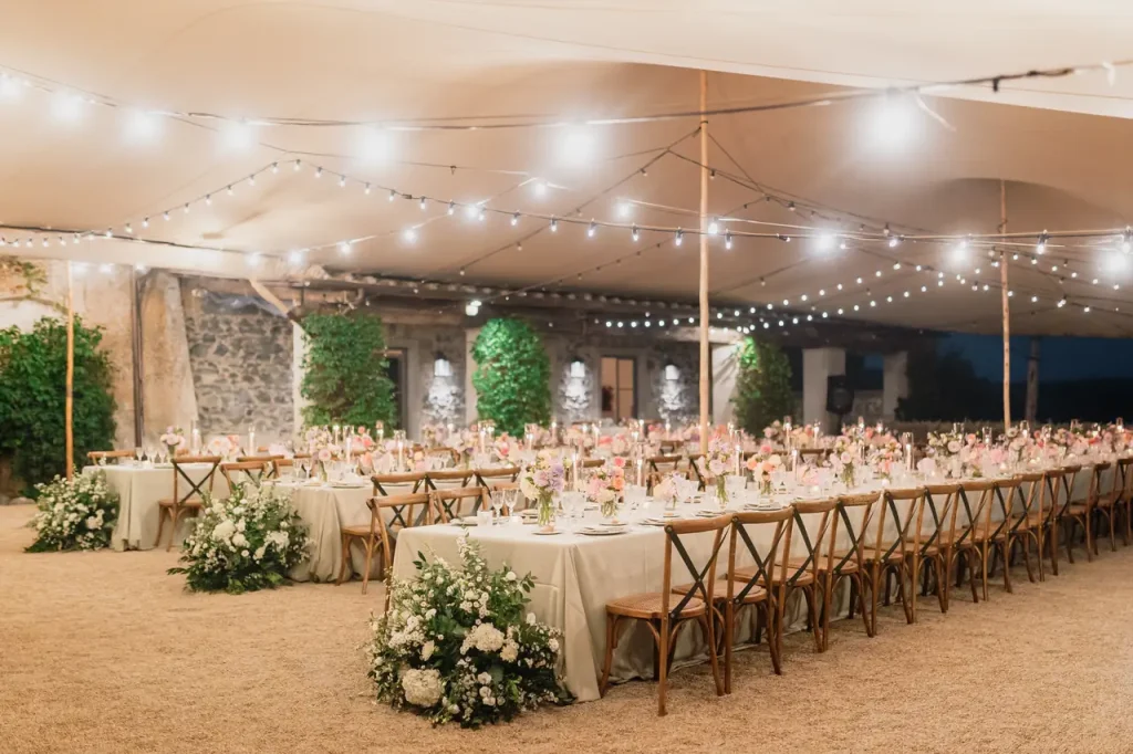 The outdoor wedding reception at casale di polline featured elegant long tables surrounded by wooden chairs, adorned with pink and green flowers. As dusk fell, string lights twinkled under an enchanting canopy, creating an enchanting atmosphere perfect for memorable celebrations.
