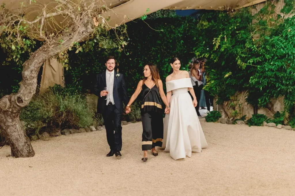 The bride and groom walk alongside wedding planner simona celani on a gravel path surrounded by greenery.