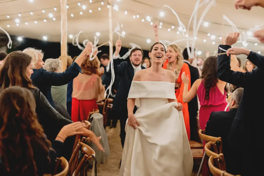 A bride walks among tables of festive guests under string lights during an outdoor event. She wears a dress with bare shoulders and holds the skirt while guests wave ribbons in celebration.