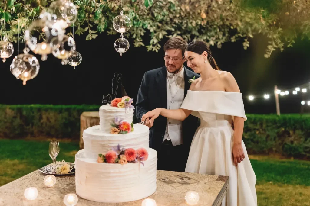A couple in formal dress cuts a three-tiered wedding cake decorated with flowers. They are standing outdoors under suspended lights.