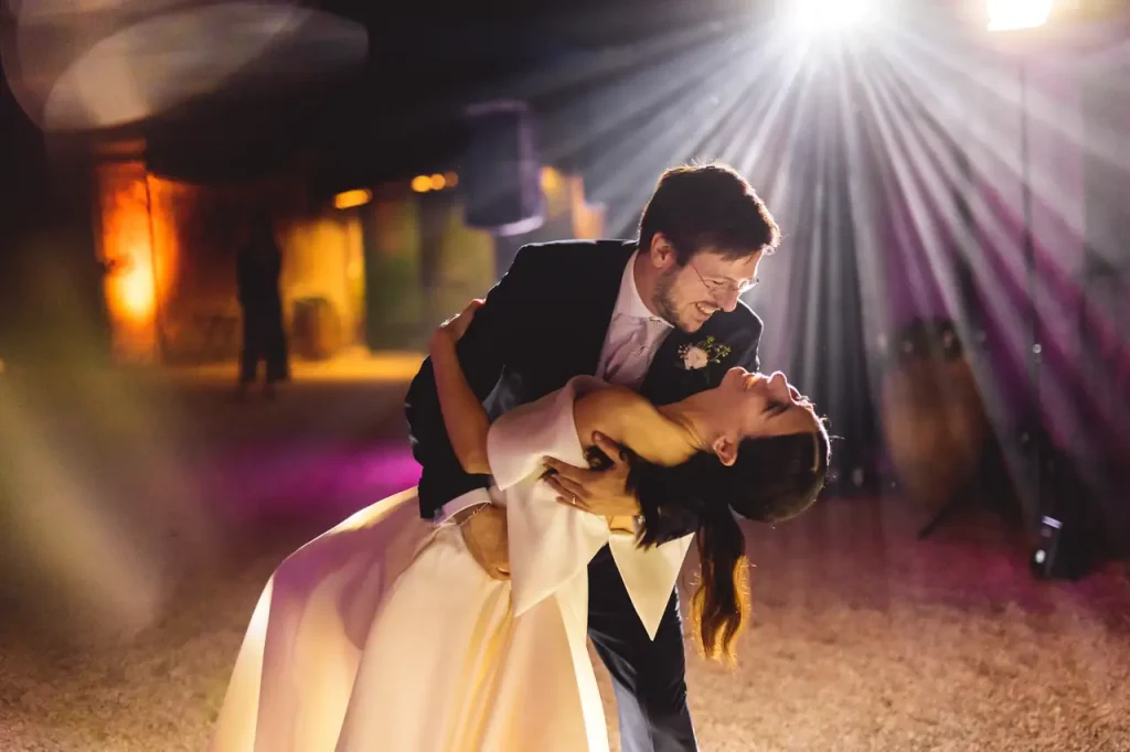 A couple dances under colored lights at an indoor event. The man accompanies the woman in a , both smiling, surrounded by a warm and vibrant atmosphere.