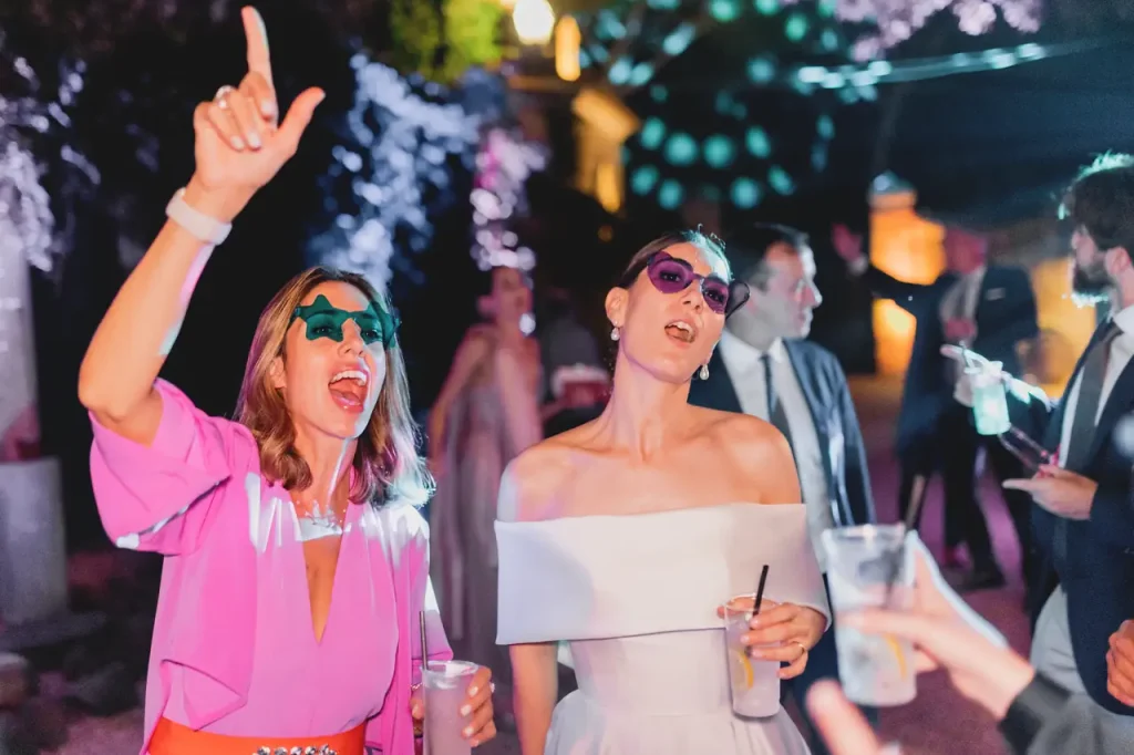 Two people wearing sunglasses sing and dance during a lively outdoor party, holding drinks.