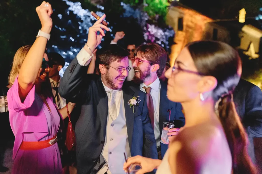 A group of people celebrate a late-night event, with two men in suits smiling and holding drinks, and a woman in the foreground with glasses.
