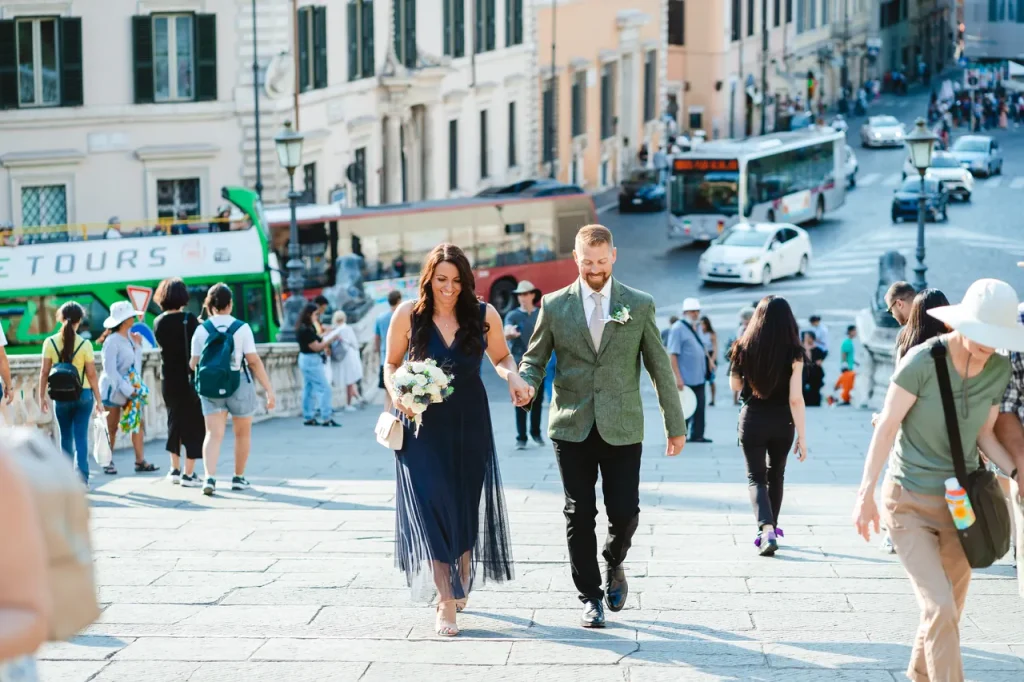 A smartly dressed couple hold hands and walk up the steps of a building in a bustling urban setting, with people around and vehicles in the background.