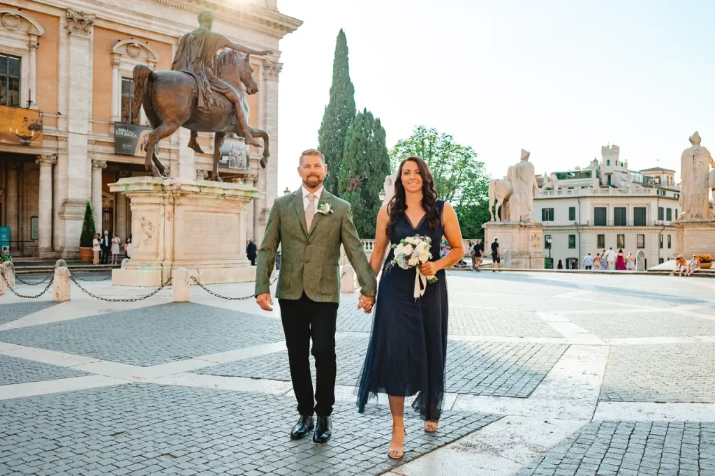 A couple holding hands is outdoors in a historic square, where the bronze equestrian statue of marcus aurelius graces the background. The man elegantly wears a suit, while the woman, holding a bouquet, wears a dark dress amid the timeless architecture.