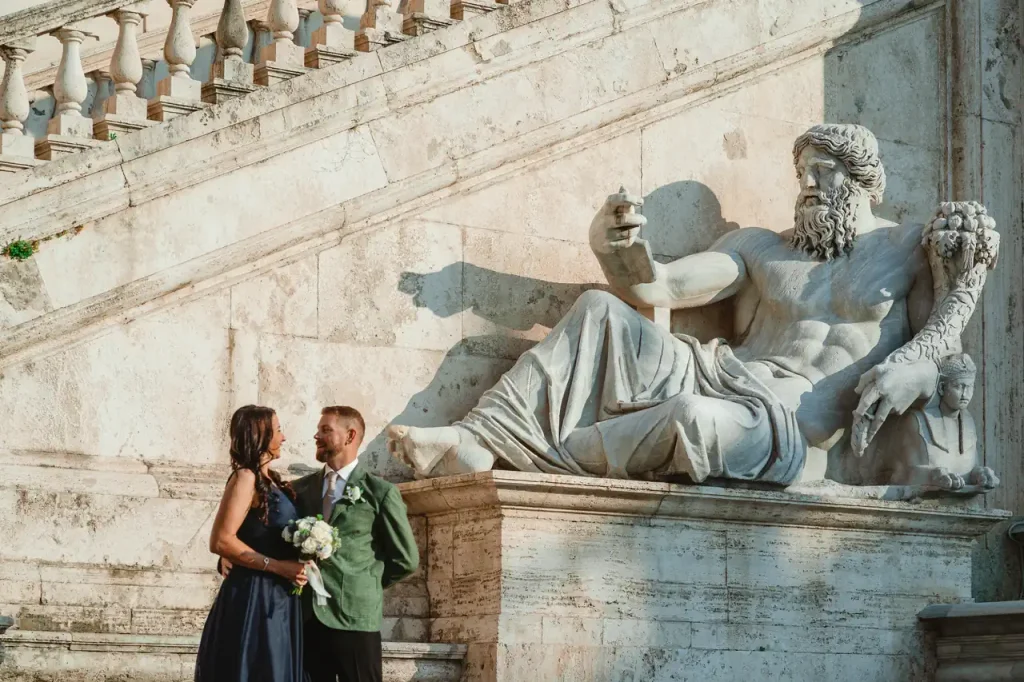 A couple dressed in formal attire are standing next to a large statue lying on a stone staircase, with the woman holding a bouquet of flowers.