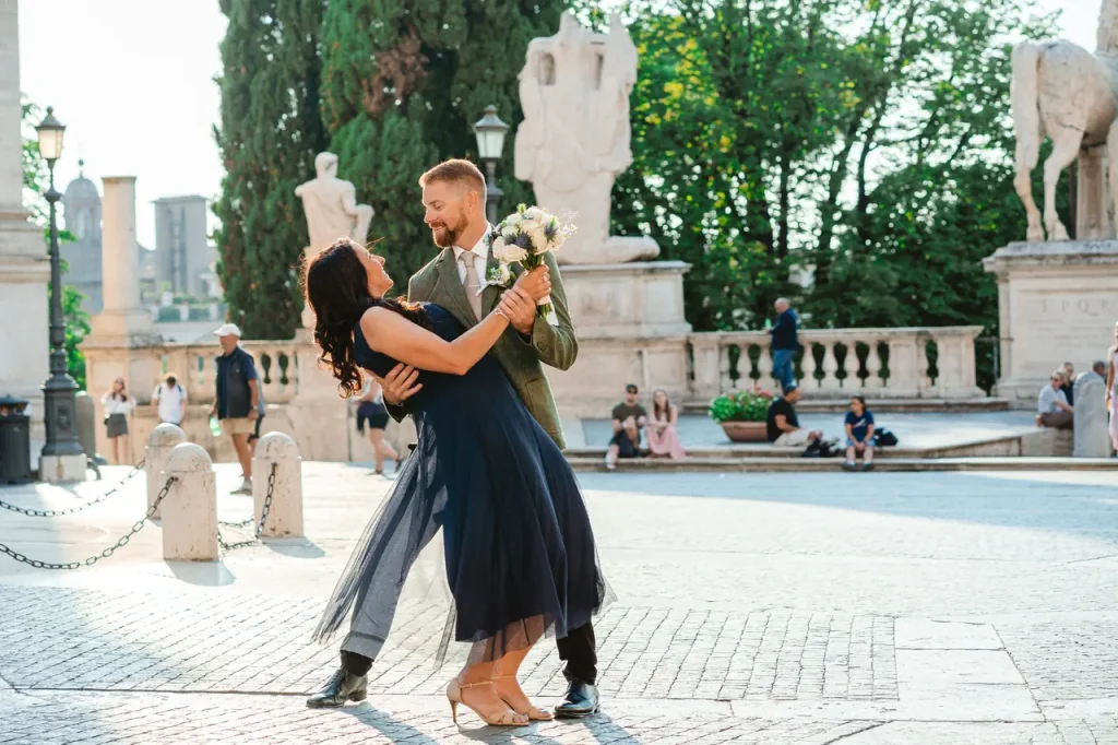 A couple dances on a cobblestone street surrounded by sculptures and vegetation. The woman wears a blue dress and the man a suit, holding a bouquet of flowers.