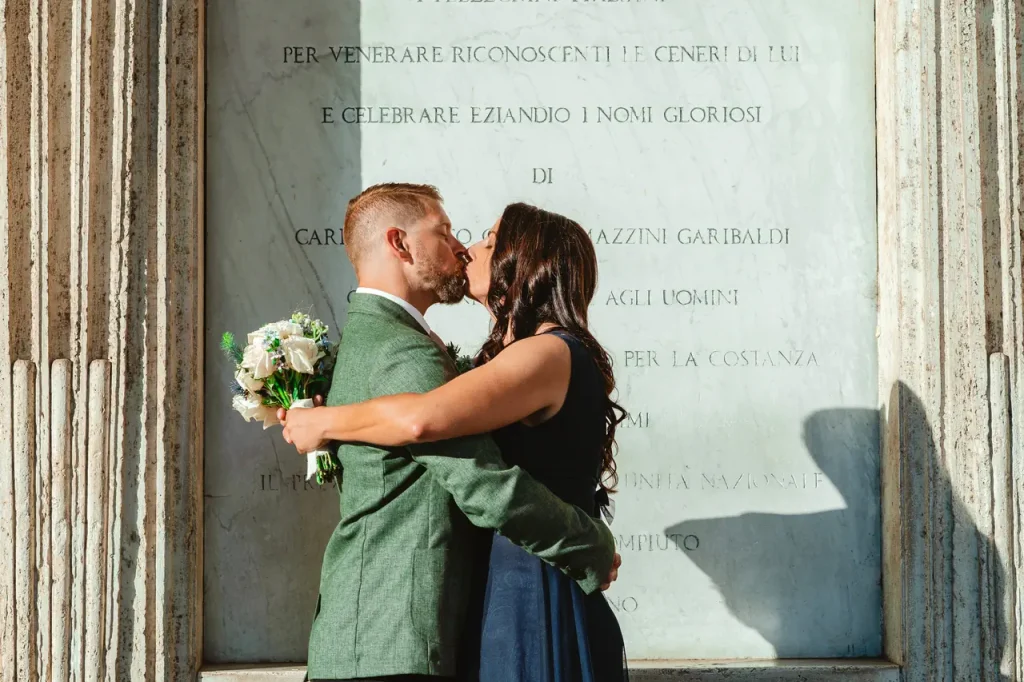A couple kiss in front of a stone wall, with the woman holding a bouquet of white flowers. The wall has engraved text, and they cast shadows on the surface.