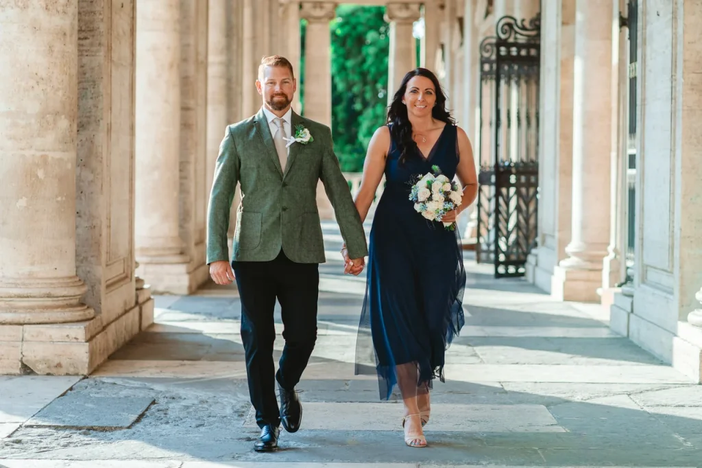 A couple dressed in formal attire walk hand in hand along a colonnaded path. The woman holds a bouquet.