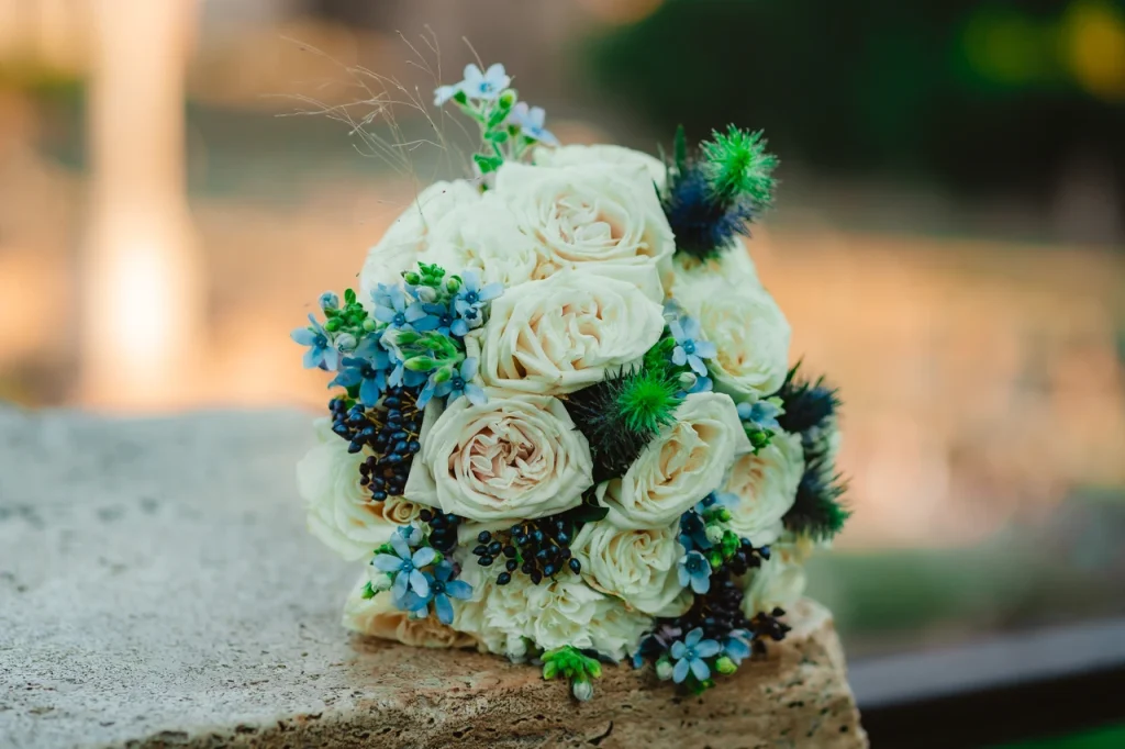A bouquet of white roses, blue flowers and pointed green details resting on a stone surface.